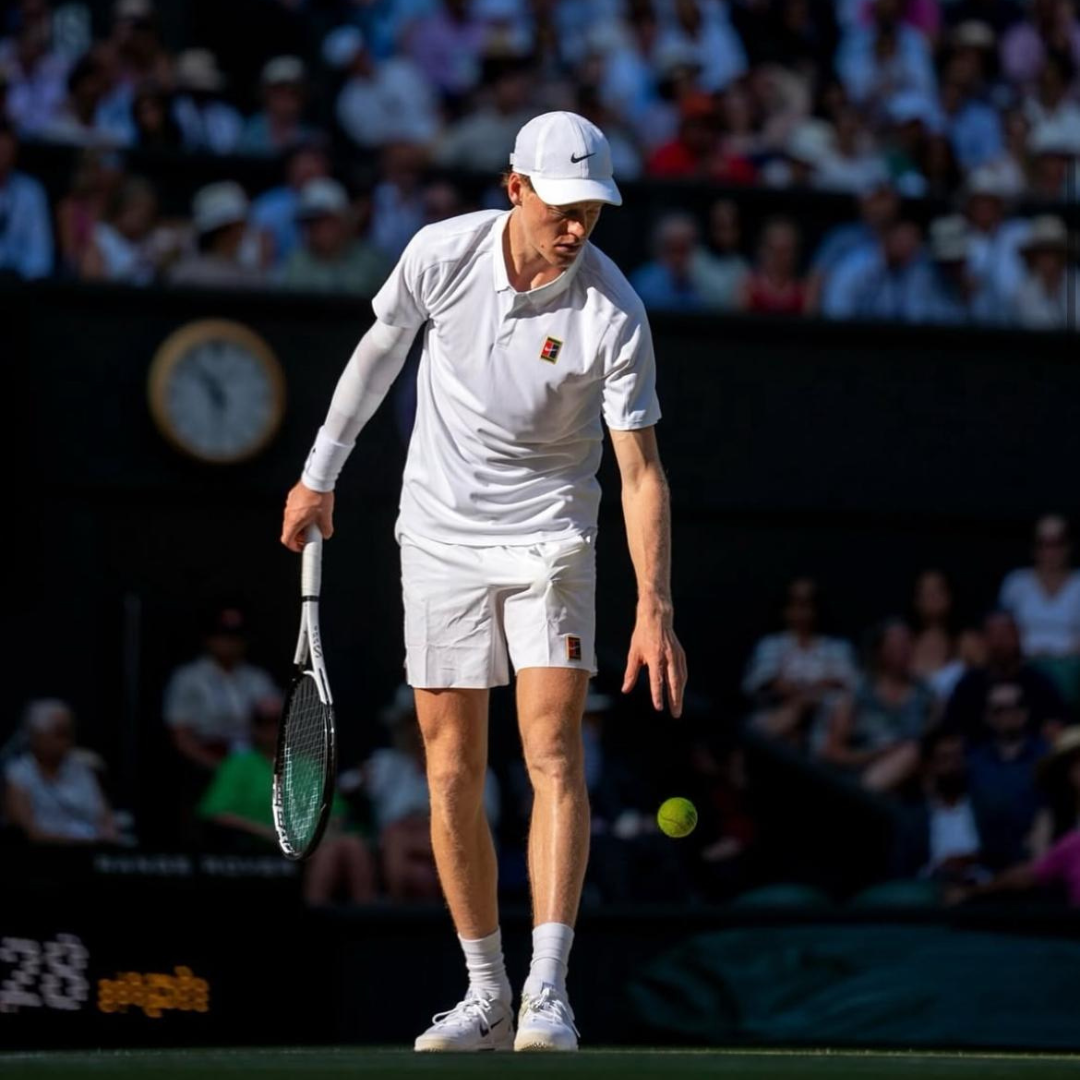 Tennis player in white outfit holding a racket and ball on a tennis court with spectators in the background.