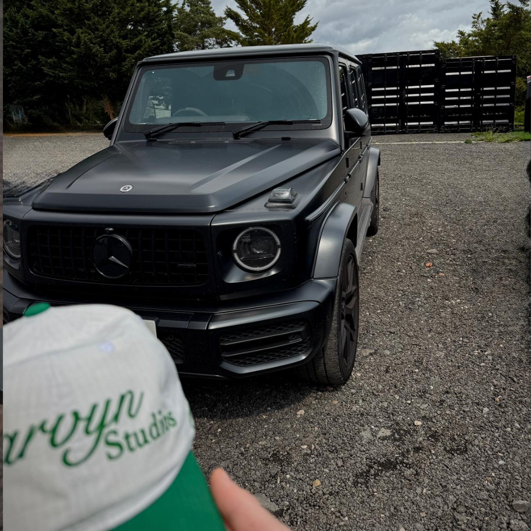 Black Mercedes-Benz G-Class SUV parked on a gravel road with a person holding a cap in the foreground.